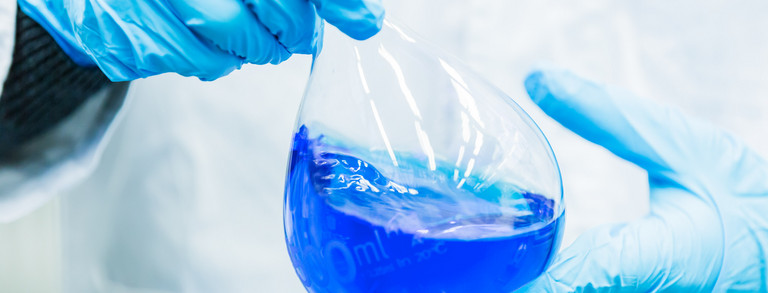 Zwei Hände in blauen Gummihandschuhen halten einen Messkolben, der mit blauer Flüssigkeit gefüllt ist.